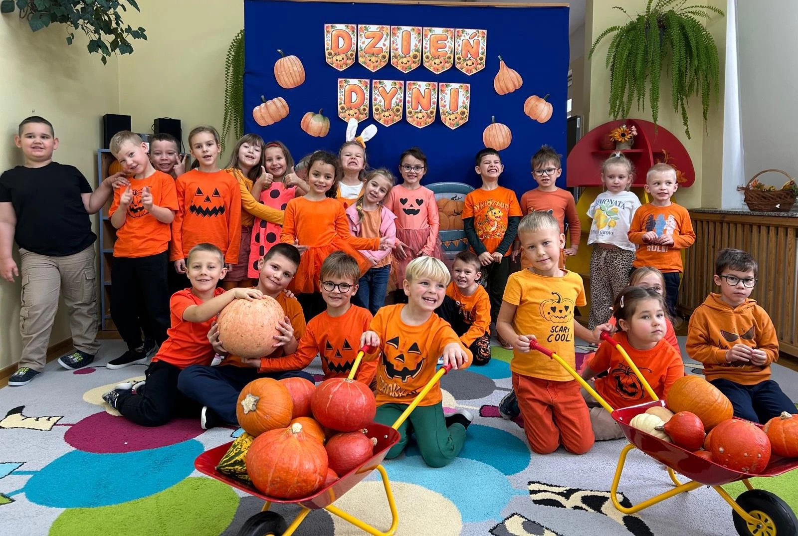 Dzieci w pomarańczowych strojach z dyniami na tle napisu „Dzień Dyni".