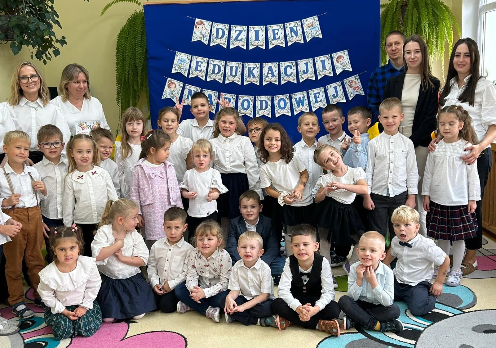 Grupa dzieci i nauczycieli pozuje na tle napisu Dzień Edukacji Narodowej.
